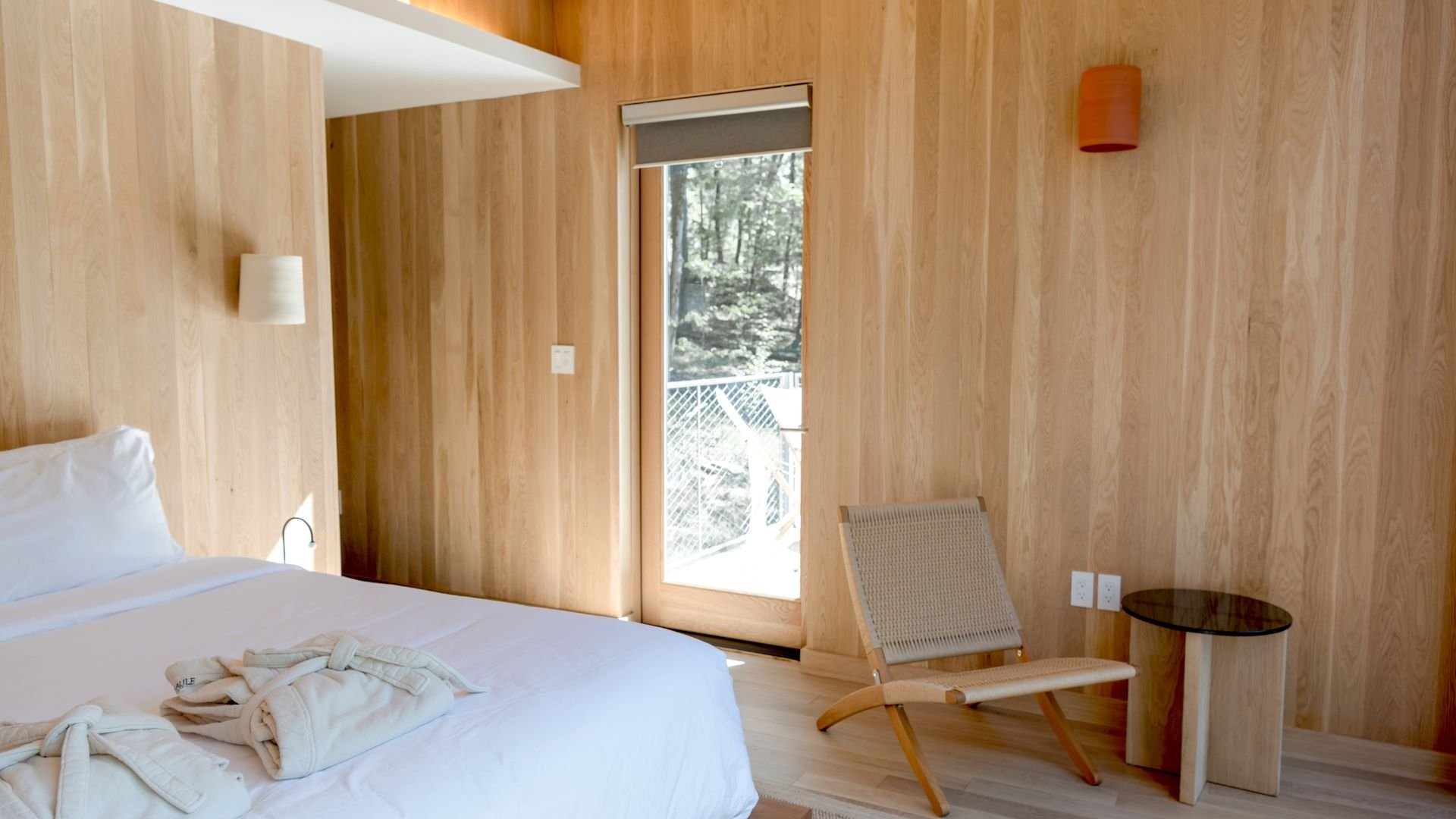 Minimalist wooden bedroom with large window overlooking forest and modern chair
